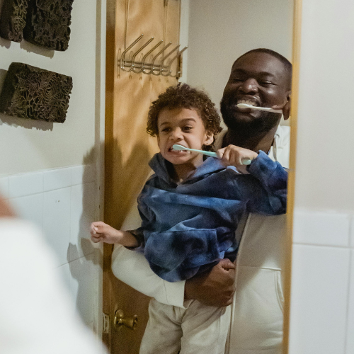 A father and his son brushing their teeth together in the mirror.